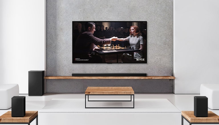 A set of 2 rear speakers, subwoofer, and a soundbar, and TV are in a white living room.  A woman and a man are playing chess on TV screen.