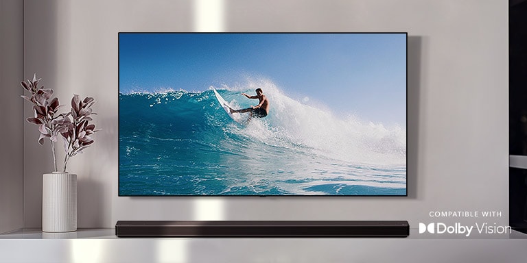 TV is on the wall. TV shows a man surfing on big wave. LG Soundbar is right below TV on a white shelf. There is a vase with a flower right next to the soundbar. (play the video)