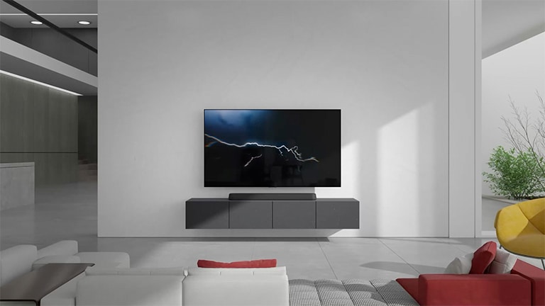 The sound bar is placed on the gray cabinet in the center and the connected TV is hung on the white wall right above, with lightnening image on it. White and red colored long sofa is placed facing the TV and the sun light is coming from the right side where a green plant is placed.