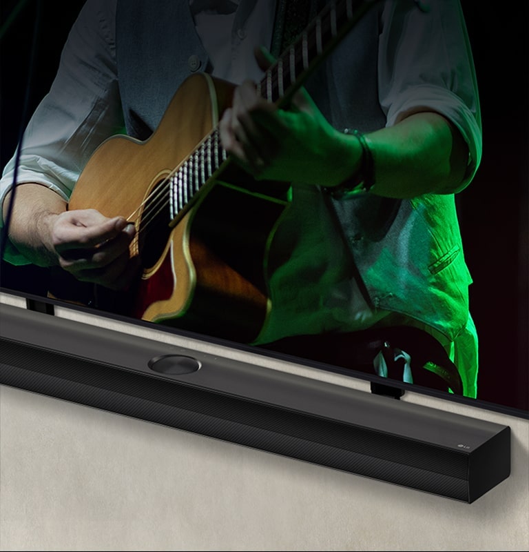LG Soundbar against a black backdrop reveals its design starting from the left corner, then pans out to show the whole soundbar. An LG QNED TV appears with Synergy Bracket. The Soundbar sits on top of the Synergy Bracket, presses against the wall with the lower screen of the TV being visible, displaying a man playing the guitar.