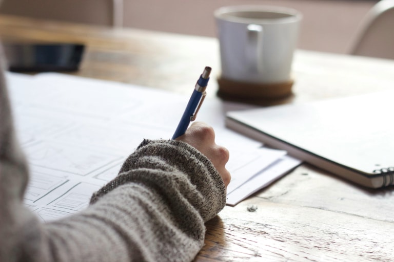 Person wearing a sweater writing on paper on a wooden table with a coffee cup in the background