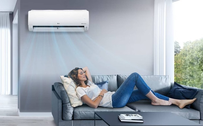 : A woman relaxing on a gray sofa in a modern living room, enjoying cool air from an LG Dual Inverter air conditioner mounted on the wall