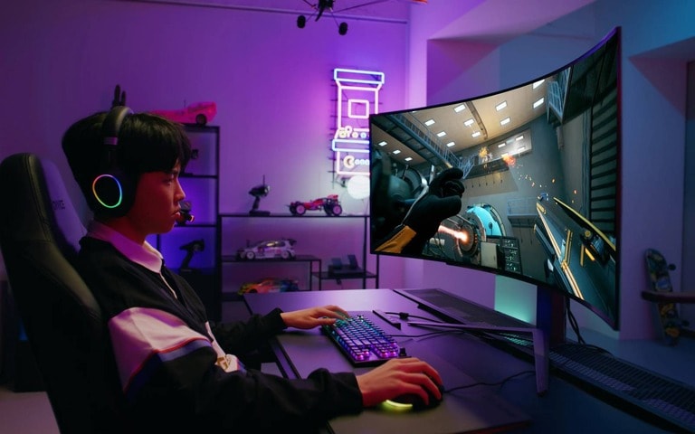 A young gamer wearing a headset plays on a curved gaming monitor, surrounded by colourful neon lights and model cars displayed on shelves in a futuristic-themed gaming room.