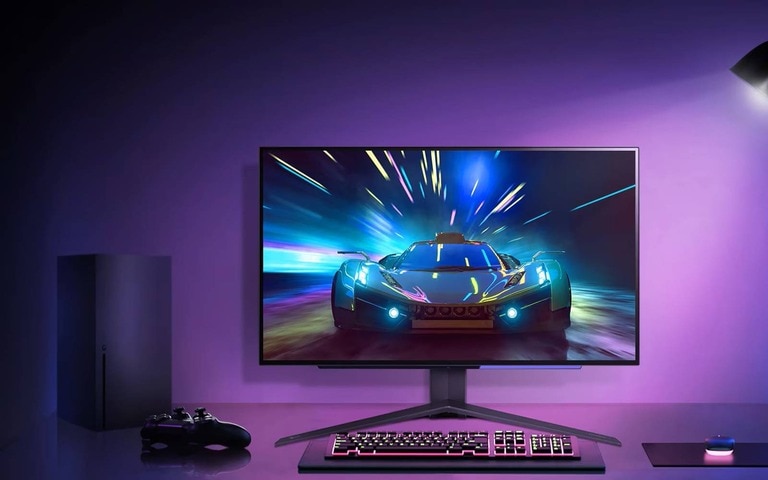 A minimalist gaming desk setup featuring a monitor displaying a high-speed car racing game, illuminated by purple and blue ambient lighting with a console controller and accessories neatly arranged.
