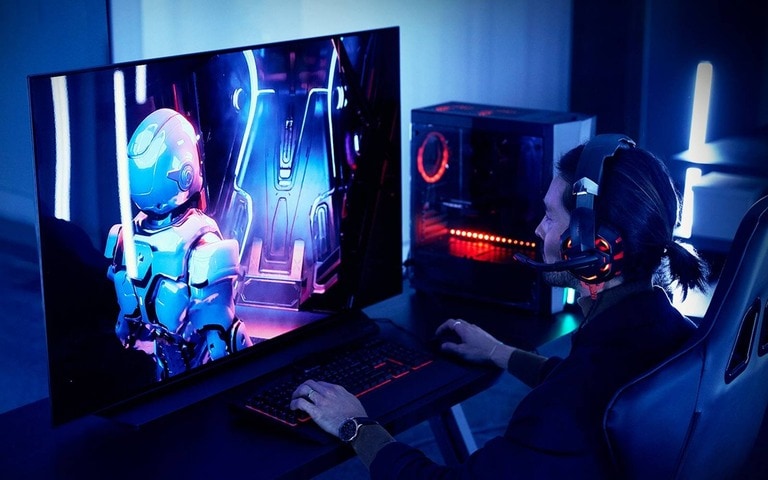 A focused gamer wearing a headset plays an intense sci-fi game on a large monitor, with a glowing custom PC setup in a dimly lit room bathed in blue and red neon lighting.