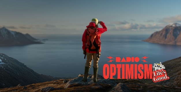 A woman wearing a red hiking jacket is standing in a place where the vast ocean and snow-covered mountains can be seen. The image also features the "Radio Optimism" slogan logo and the "Life's Good." logo alongside the woman.