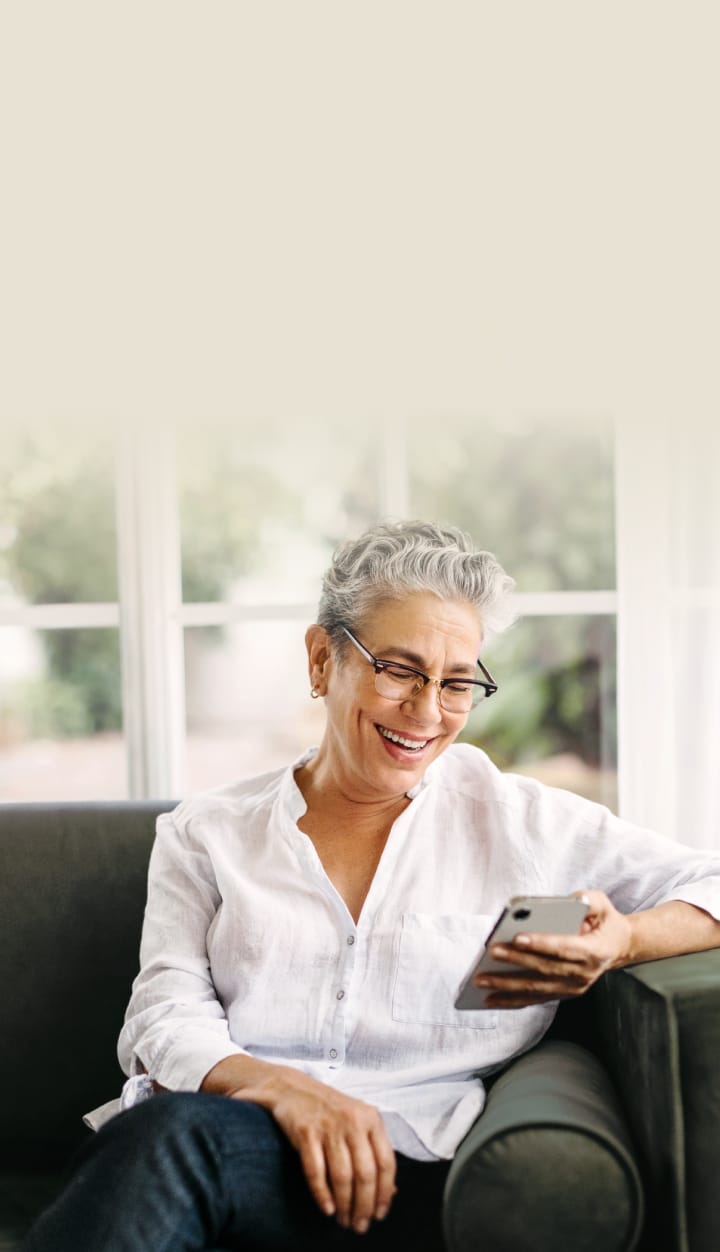 An elderly woman with gray hair, wearing glasses, sitting on a couch and smiling while using a phone.	