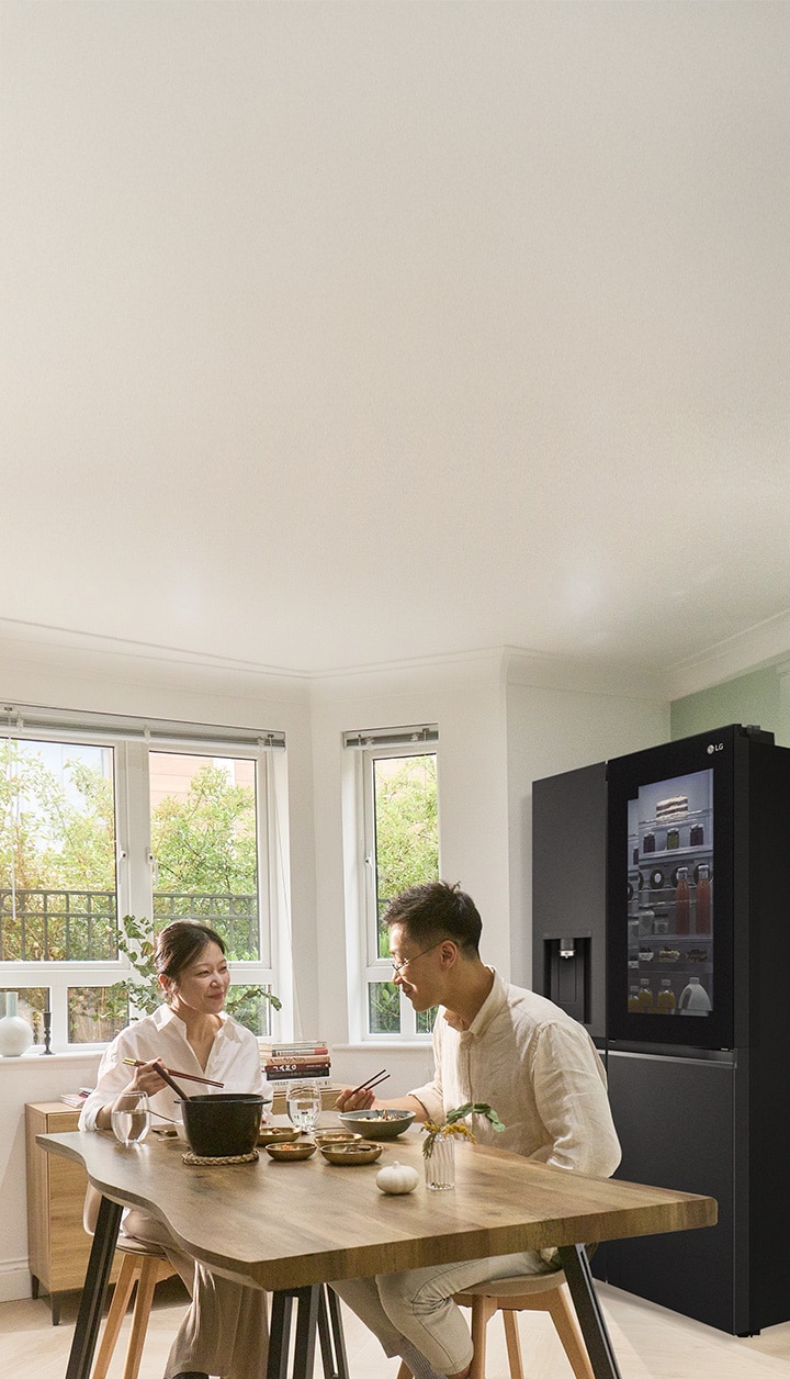 A couple sitting at a dining table having a meal with a refrigerator beside them