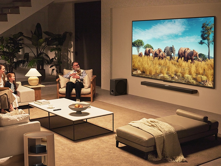 A family sits on a couch facing a LG QNED TV mounted on a wall above a LG Soundbar in a modern living room space.  Zero Connect Box sits inside a coffee table.