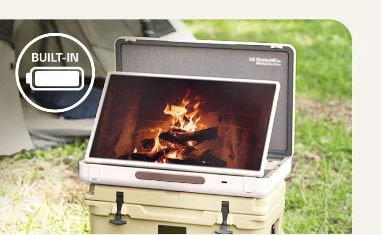 The LG StanbyME Go is placed in front of a tent, and the screen displays one of the relaxing themes--a fireplace. On the left top corner, built-in battery icon is shown.