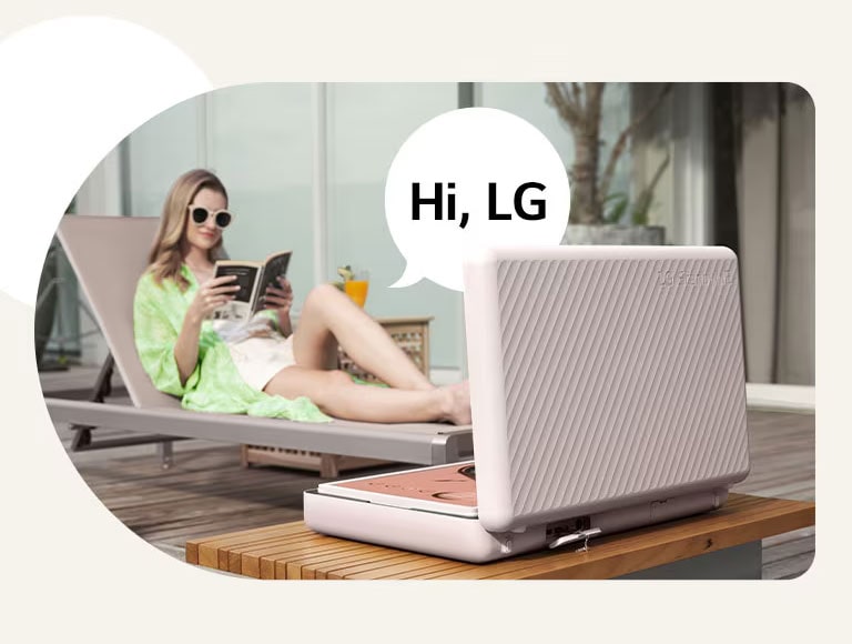 Rear view of the LG StanbyME Go, and it's placed right in front of the patio table. A woman is chilling out on the beach chair, control the screen with her voice. To illustrate this, a speech bubble with Hi, LG text is shown on the right side of her.