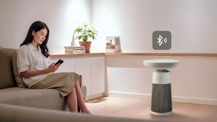 Hình ảnh một người phụ nữ đang sử dụng điện thoại thông minh có loa LG Aerospeaker trong phòng khách, hiển thị logo Bluetooth.