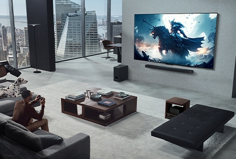 Phòng khách với một người đang chơi trò chơi điện tử trên TV LG True Wireless và LG Soundbar treo tường. Anh cầm một bộ điều khiển trong tay, nhưng máy chơi game không dễ để nhìn thấy ngay lập tức. Cũng không có dây nào có thể nhìn thấy. Hộp kết nối không dây được giấu kín, hầu như không nhìn thấy bên dưới bàn phụ.