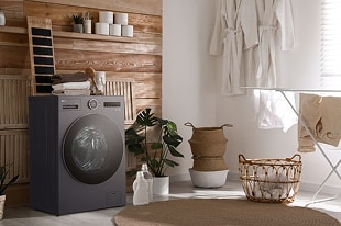 Máy giặt sấy LG thế hệ mới với thiết kế hiện đại đặt trong phòng giặt tông gỗ ấm áp, kèm theo giỏ đựng đồ, cây xanh và giá phơi đồ, tạo cảm giác tiện nghi và tinh tế.