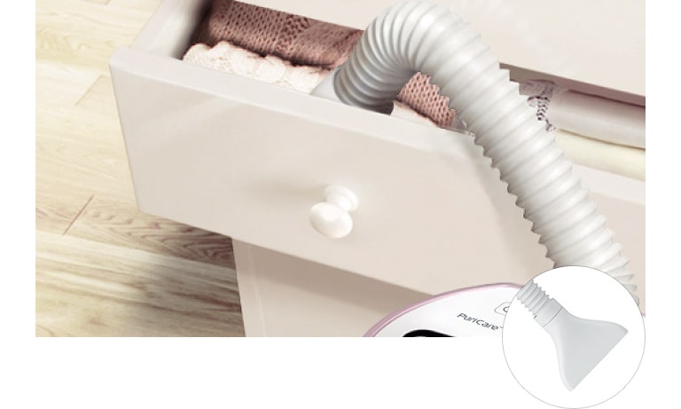 A flexible vacuum hose extends into an open drawer filled with neatly folded clothes on a light wooden floor.