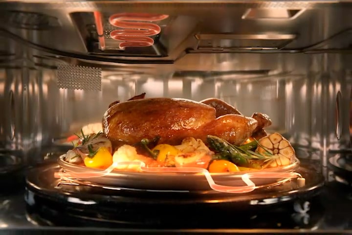 Whole chicken roasting on a rotating tray inside an LG SolarDOM, evenly cooked with surrounding vegetables.