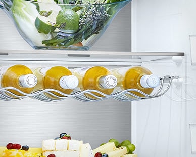 Five beverage bottles are placed in the wine rack that hangs under a shelf inside the fridge with other produce on the shelves surrounding.