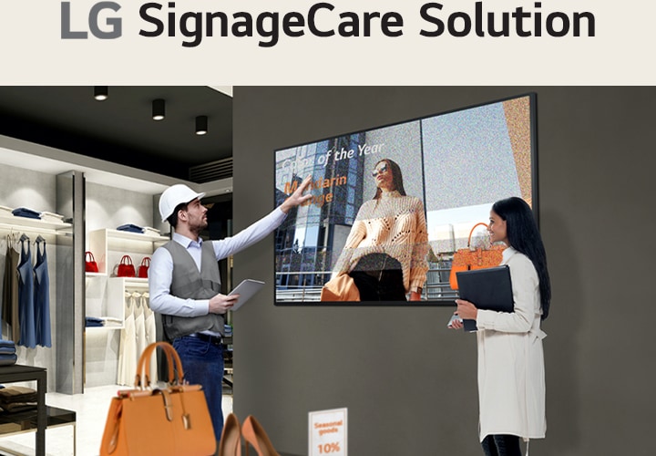 A retail store employee in a vest and hard hat gestures toward a digital display showing a woman with "Employee of the Year" text. Another person, holding a folder, observes the screen in a modern retail space with shelves of clothing and bags.