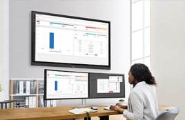 Woman working at a desk in an office, analyzing data displayed on multiple monitors including a large wall-mounted screen.