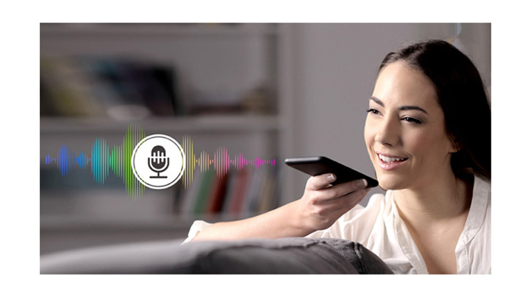 A woman is controlling TV by speaking to a remote control which has voice recognition.
