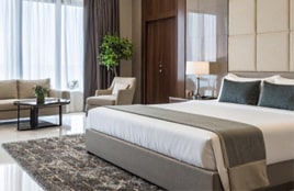 Interior moderno de una habitación de hotel con una cama perfectamente tendida, iluminación suave, sillones y grandes ventanas con luz natural.
