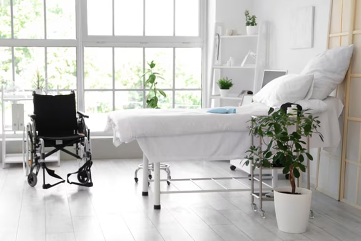 Habitación de sala de atención médica con cama, silla de ruedas y ventanas amplias