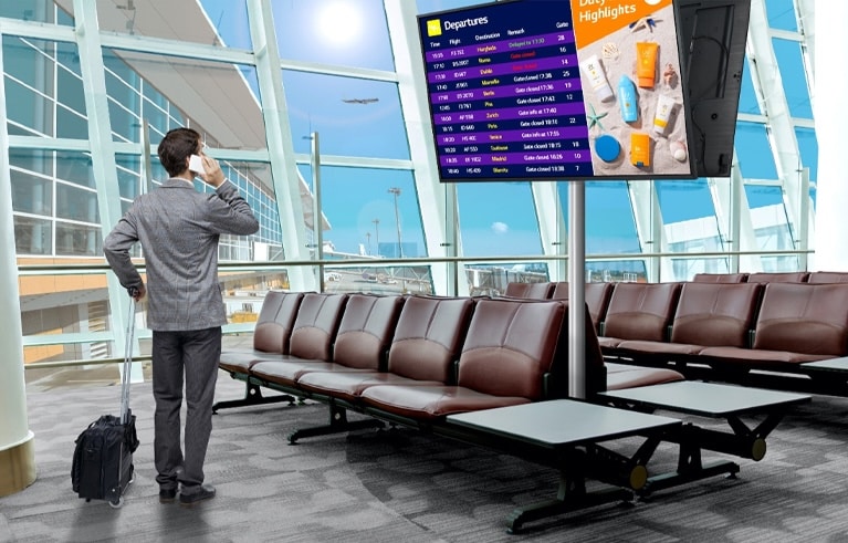 Un viajero se encuentra en la sala de espera de un aeropuerto, revisando su teléfono mientras mira el panel digital que muestra los horarios de salida de los vuelos. La escena destaca el uso moderno y eficiente de la señalización para proporcionar información actualizada en tiempo real.
