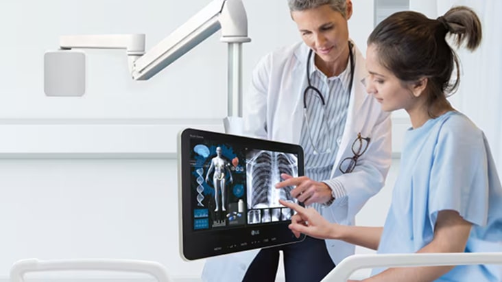 Un médico y un paciente en una habitación de hospital revisan imágenes médicas que se muestran en un monitor con pantalla táctil junto a la cama del paciente.