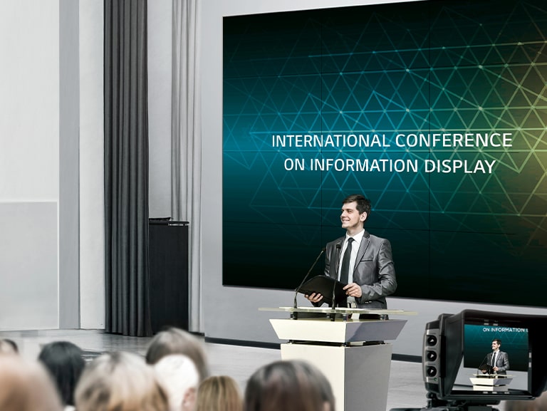 Un grand mur d'images est installé dans la salle de conférence et un homme tient une conférence devant de nombreuses personnes.