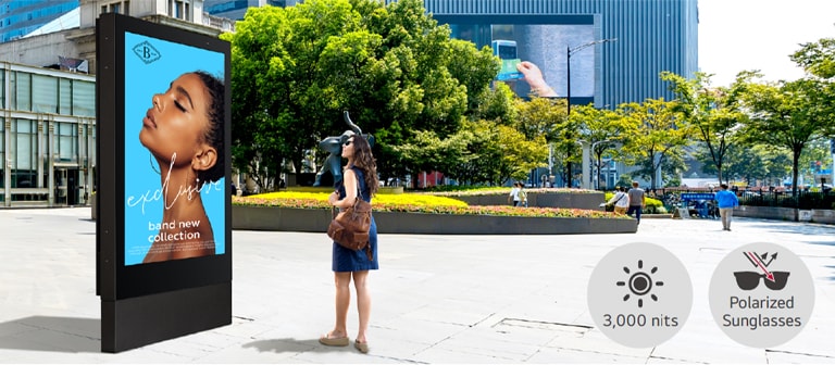 A large-sized display is installed on the street, and a woman is looking at a vivid-quality advertisement on the screen.