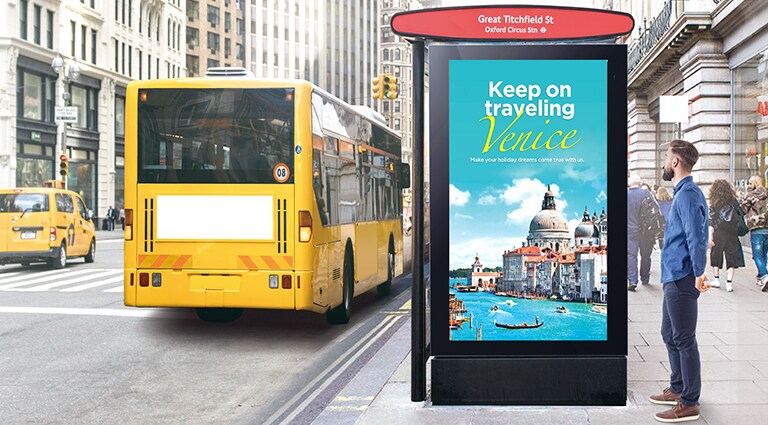 A large display is installed at the bus stop, and a man is looking at a vividly pictured quality advertisement on it.