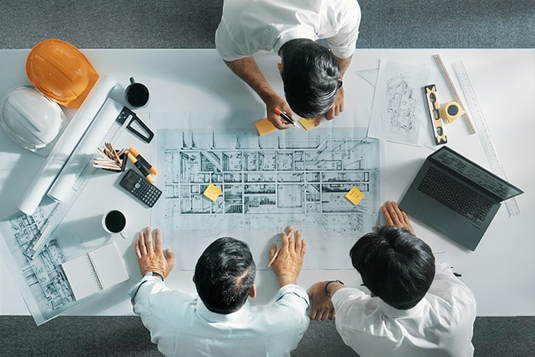 Top-down view of three engineers analyzing large blueprints on a desk, representing customer commitment.