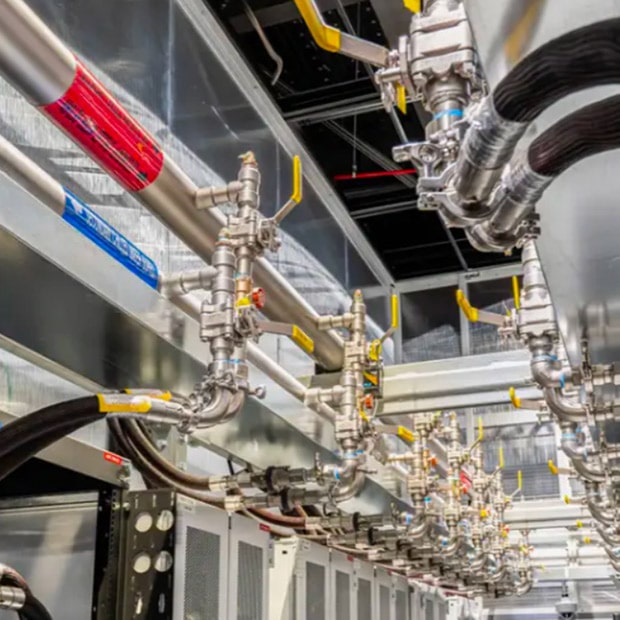 An overhead view of industrial cooling pipes, valves, and thick hoses connected to server racks in a data center.	