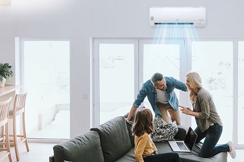 Family in a bright living room using a laptop under a wall-mounted air conditioner with cool air flowing.