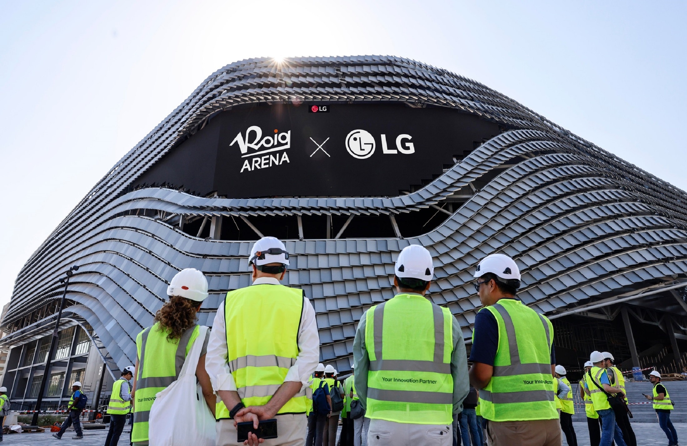 Construction workers in safety vests and helmets gathered outside Roig Arena, with a large LG × Roig Arena logo displayed on the curved modern façade