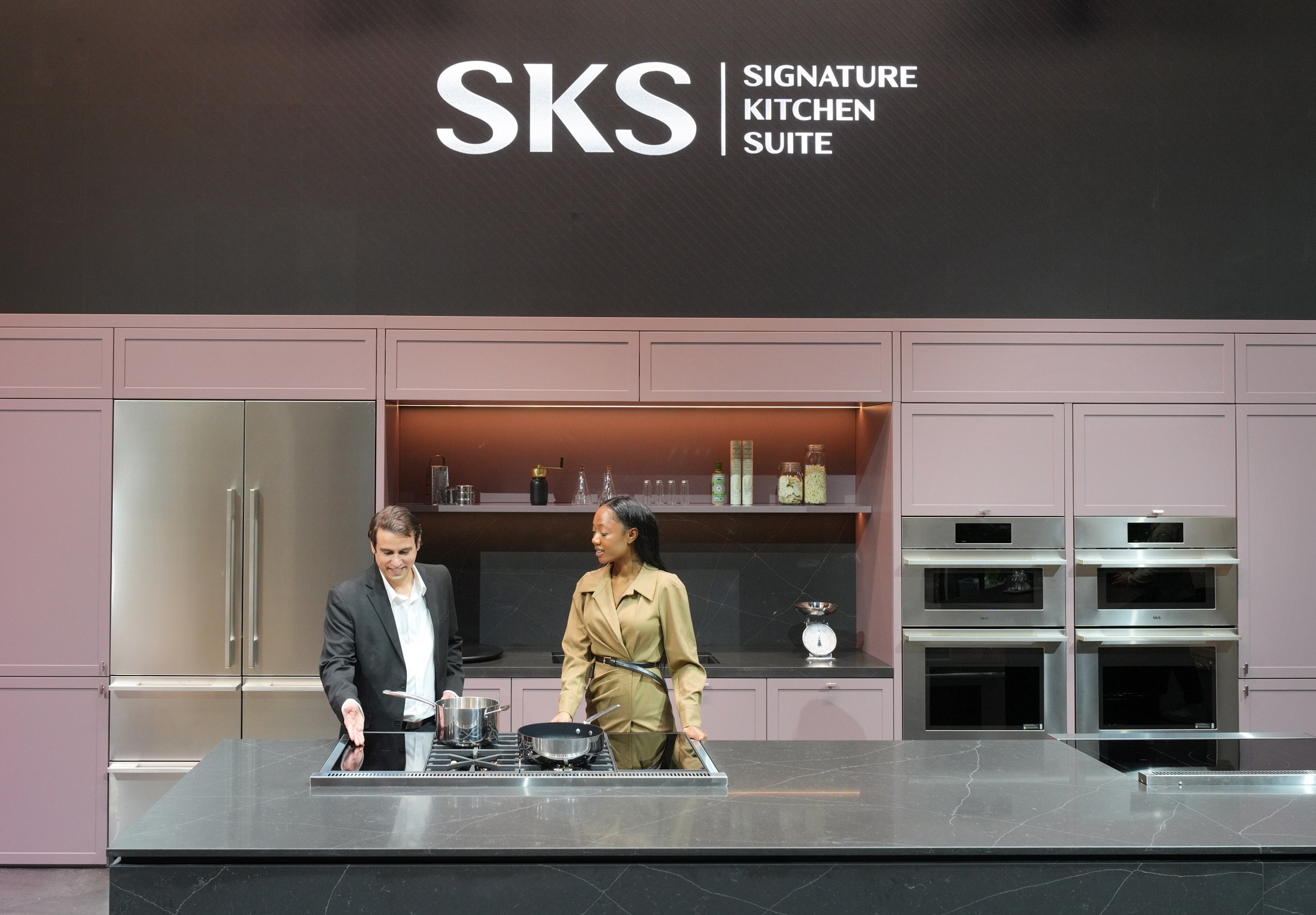 Two people standing in a modern kitchen showroom under the logo 'SKS | Signature Kitchen Suite', demonstrating built-in appliances including a refrigerator, ovens, and stovetop.