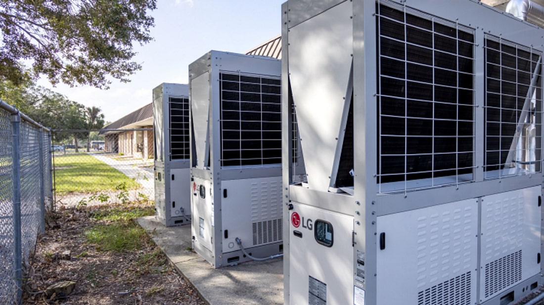 Outdoor installation of multiple large LG industrial HVAC units lined up on a concrete base, adjacent to a fenced area and building