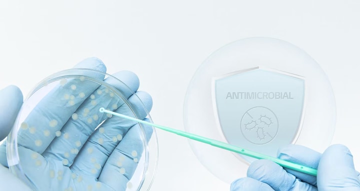 Gloved hands holding a petri dish with bacterial colonies while using a swab, with an antimicrobial shield icon in the background.