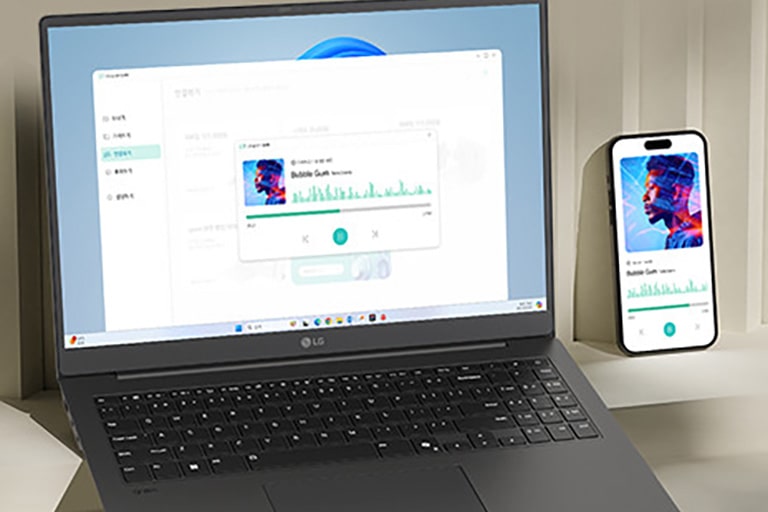 A laptop and a smartphone are placed on a desk, both displaying the same music playback interface, indicating synchronized functionality. The setup highlights seamless integration between devices for multimedia activities.