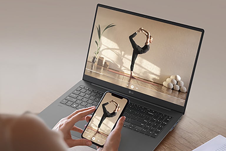 The image features a person holding a smartphone in front of a gray laptop, both displaying the same video of a woman performing a yoga pose. The scene highlights seamless screen mirroring or content sharing between devices.