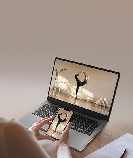 The image features a person holding a smartphone in front of a gray laptop, both displaying the same video of a woman performing a yoga pose. The scene highlights seamless screen mirroring or content sharing between devices.