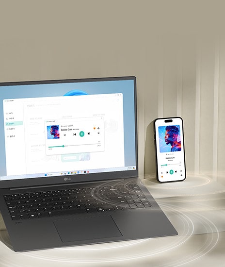 A laptop and a smartphone are placed on a desk, both displaying the same music playback interface, indicating synchronized functionality. The setup highlights seamless integration between devices for multimedia activities.