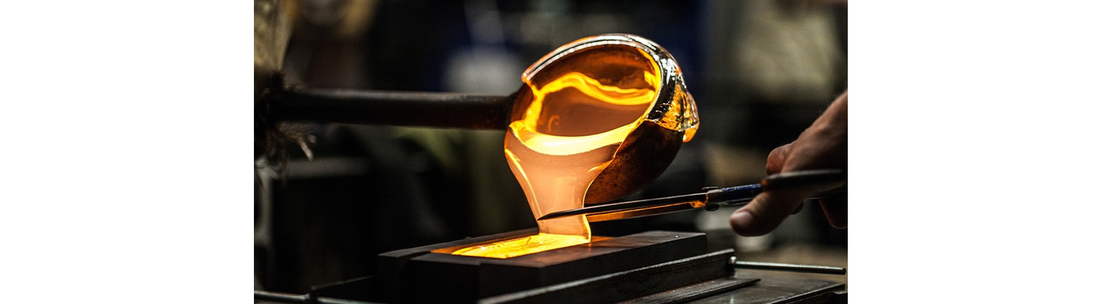 A close-up of a glassblowing process, focusing on a large blob of molten, glowing glass being worked.