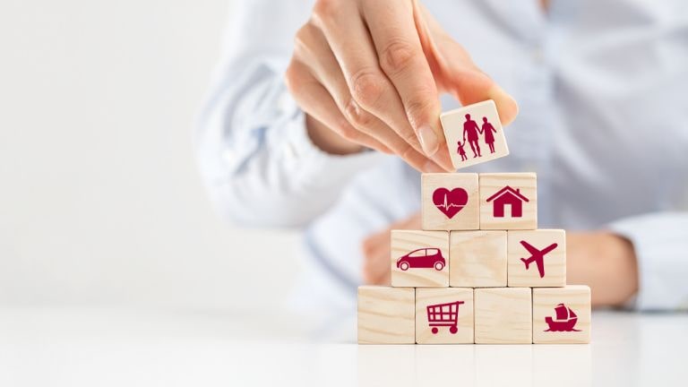 Hand stacking wooden blocks with red icons representing family, health, home, car, travel, shopping, and lifestyle.