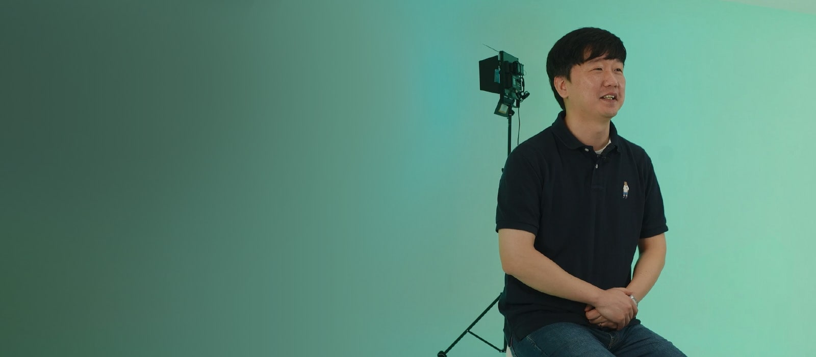 LG CAE Team Member wearing black short-sleeved shirt is sitting on a chair with green background.