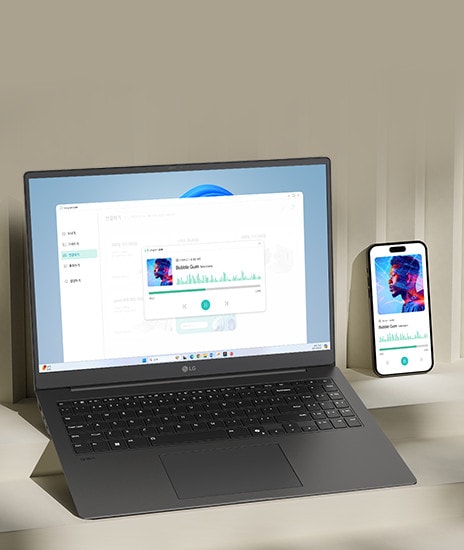 A laptop and a smartphone are placed on a desk, both displaying the same music playback interface, indicating synchronized functionality. The setup highlights seamless integration between devices for multimedia activities.