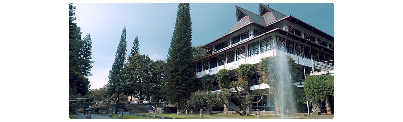 Modern campus building with triangular roofs, glass facade, fountain, and pine trees in landscaped setting