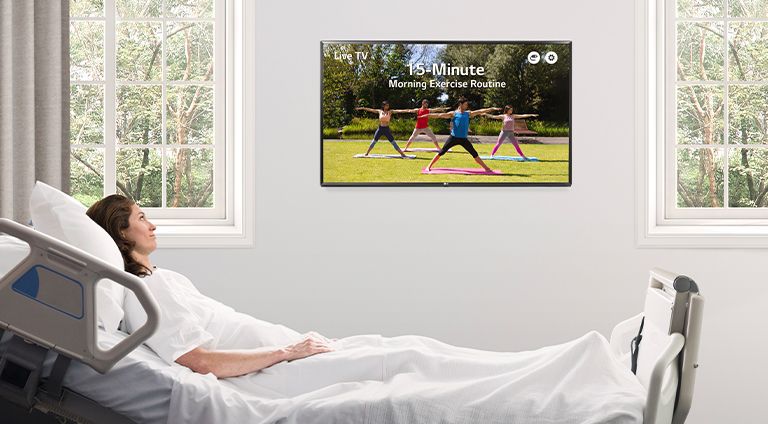The patient is lying in the hospital room’s bed and watching the TV hanging on the wall.