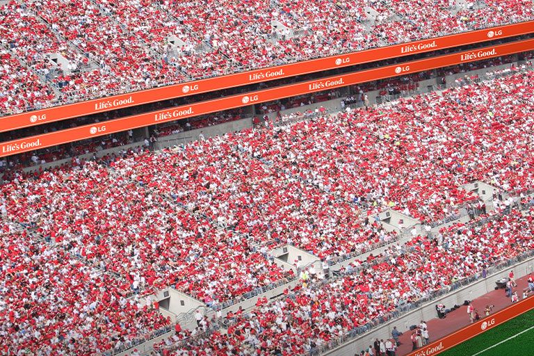 Ribbon LEDs are installed in a continuous line along the railing wall of the upper-tier stands in the soccer stadium, clearly displaying advertisements.