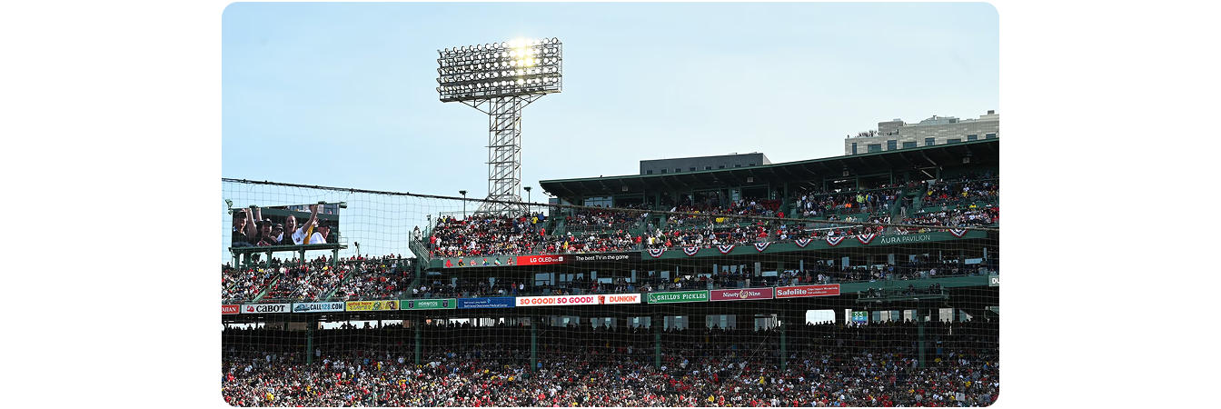 LG LED ribbon-board signage at Fenway Park displaying "LG OLED — The best TV in the game" along the grandstand façade, with packed crowd, sponsor banners and the iconic centerfield light tower above